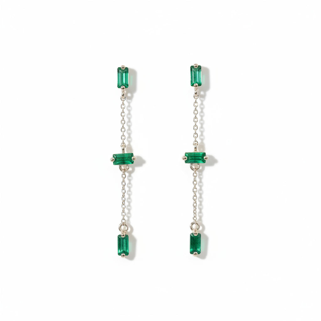 A pair of long drop earrings with rectangular green gemstones and silver-tone chains, displayed on a textured white background with a repeating 'MINIMAL ATTELER' watermark.