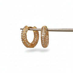 A pair of gold hoop earrings covered in small clear stones, displayed on a metal stand against a light background with a repeating MINIMAL ATELER watermark.