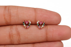 A pair of small heart-shaped stud earrings with multicolored gemstones in a gold-tone setting, shown resting on a person's fingers for scale.