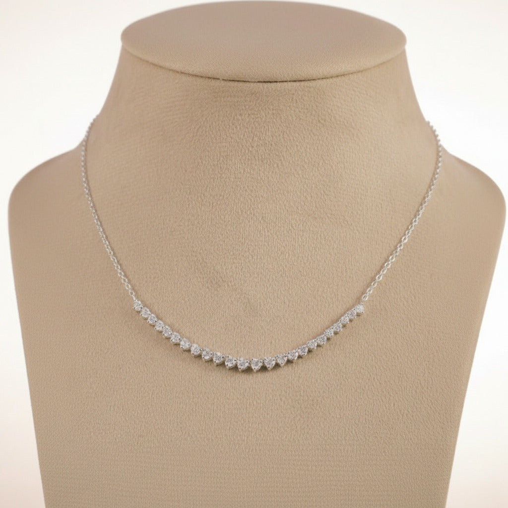 A delicate silver-toned chain necklace with a curved row of small clear stones, displayed on a beige jewelry bust against a light purple background.
