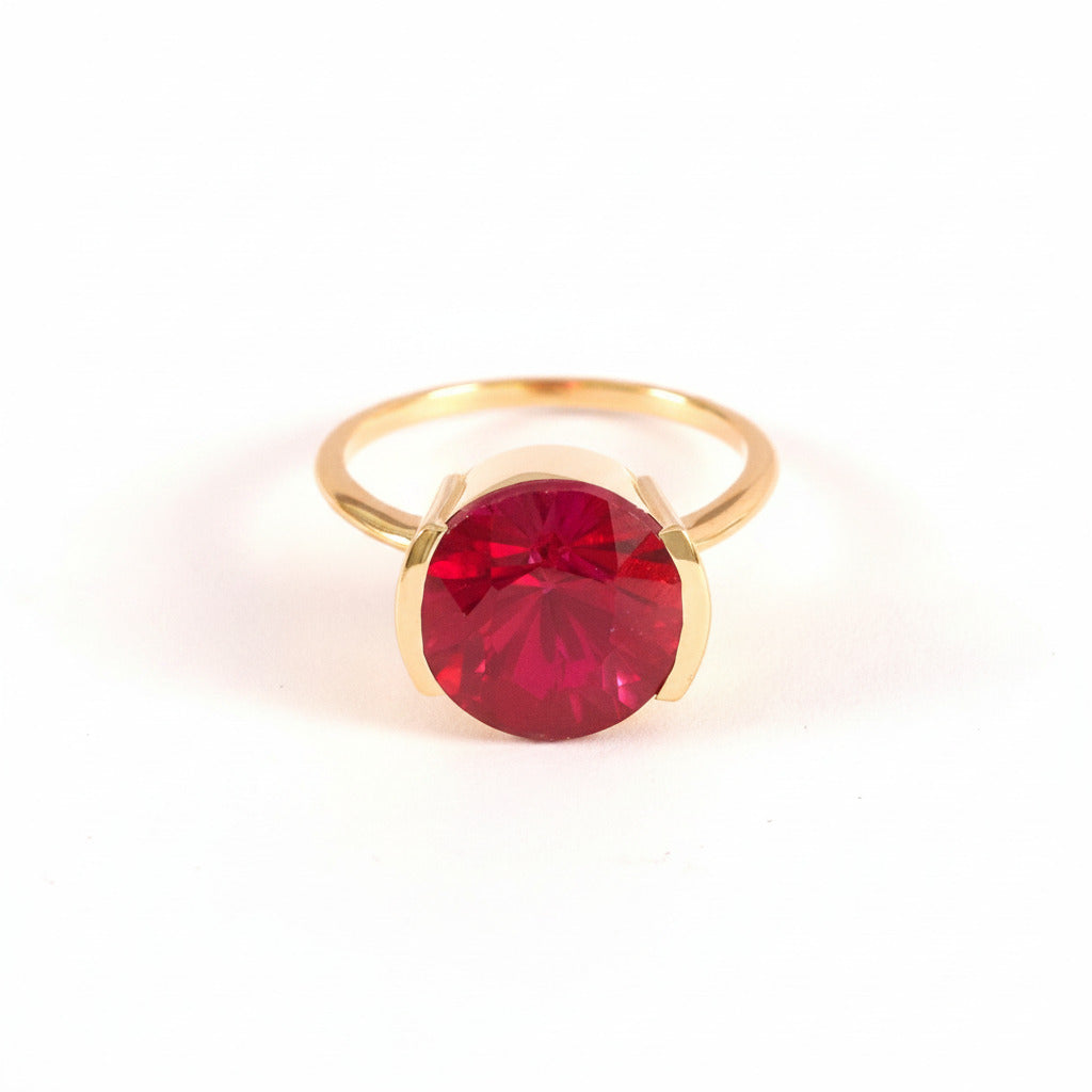 A gold ring with a large, round, deep red gemstone, photographed on a light pink background with a repeating diagonal watermark pattern.