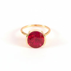 A gold ring with a large, round, deep red gemstone, photographed on a light pink background with a repeating diagonal watermark pattern.