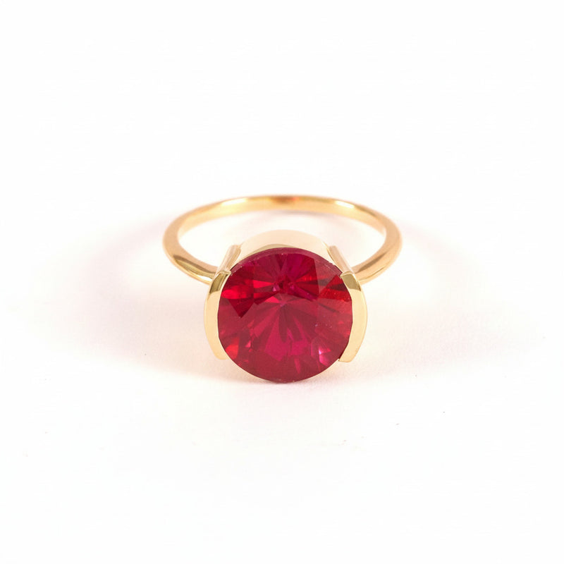 A gold ring with a large, round, deep red gemstone, photographed on a light pink background with a repeating diagonal watermark pattern.