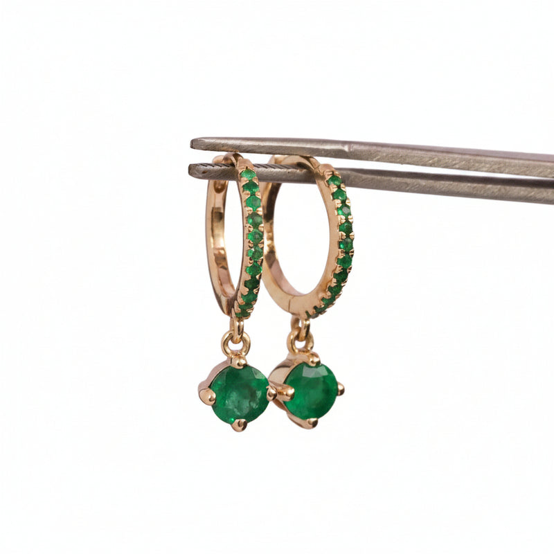 Gold huggie hoop earrings with green stone accents and dangling round green gemstones, held by metal tweezers against a pink watermarked background.