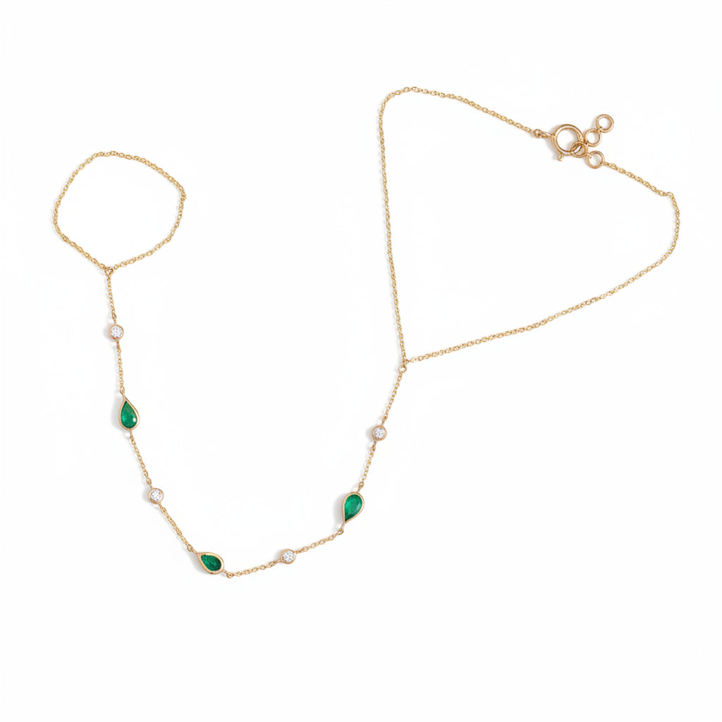 A delicate gold chain anklet featuring three green teardrop-shaped stones and several small white pearl-like beads, laid flat on a textured beige fabric background.