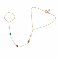 A delicate gold chain anklet featuring three green teardrop-shaped stones and several small white pearl-like beads, laid flat on a textured beige fabric background.