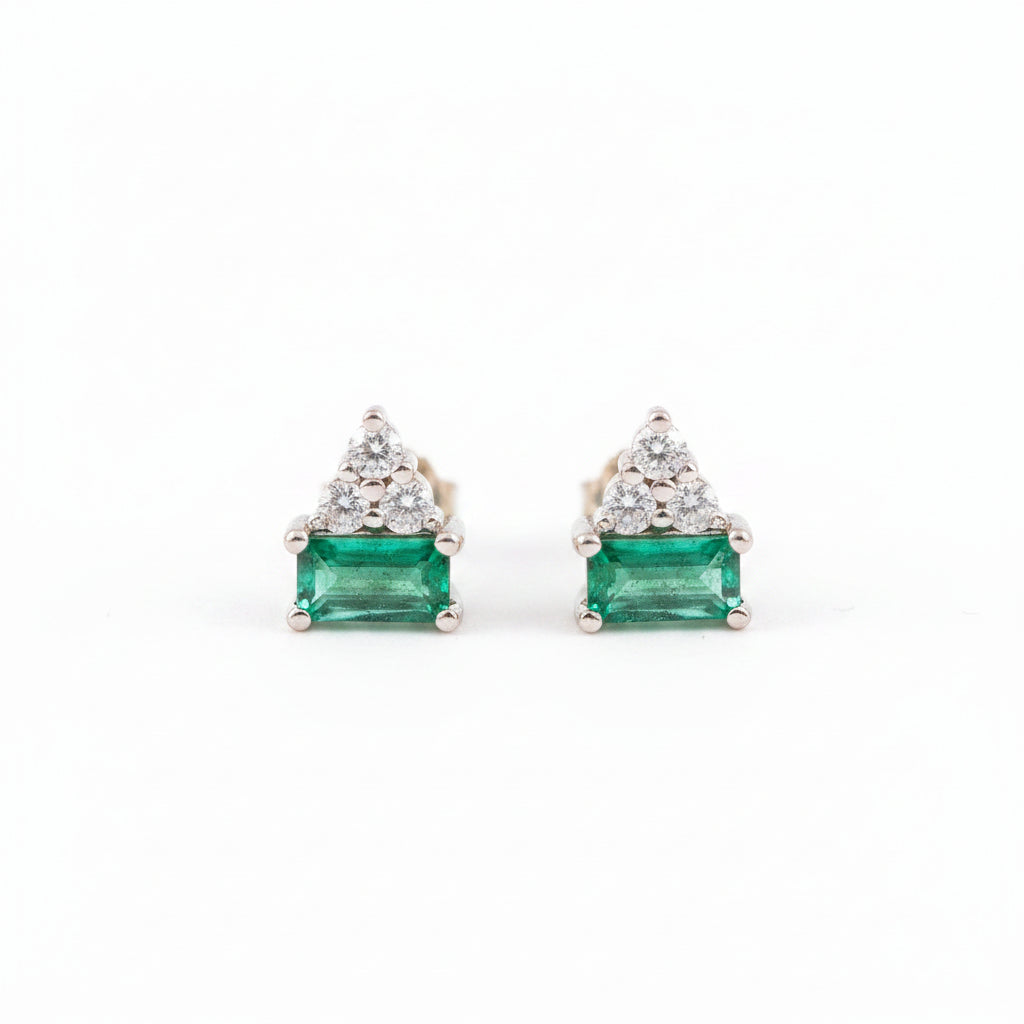 A pair of gold triangular stud earrings featuring a rectangular green emerald-cut center stone and a small cluster of clear stones above it, displayed on a textured light pink surface with a repeating 'MINIMAL ATELIER' watermark.
