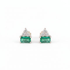 A pair of gold triangular stud earrings featuring a rectangular green emerald-cut center stone and a small cluster of clear stones above it, displayed on a textured light pink surface with a repeating 'MINIMAL ATELIER' watermark.
