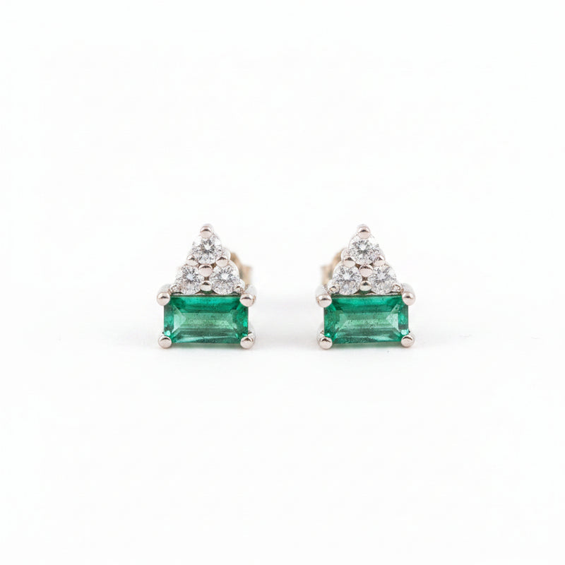 A pair of gold triangular stud earrings featuring a rectangular green emerald-cut center stone and a small cluster of clear stones above it, displayed on a textured light pink surface with a repeating 'MINIMAL ATELIER' watermark.