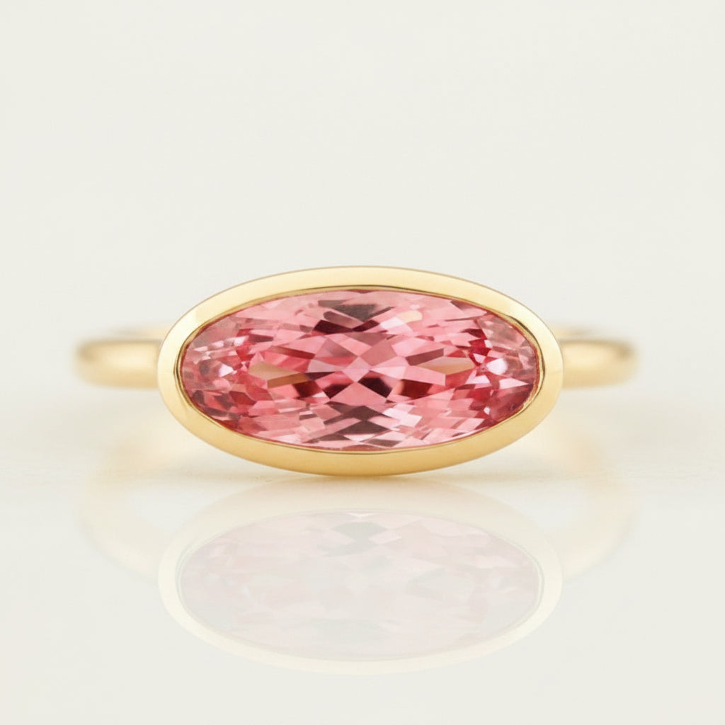 An oval-cut pink gemstone ring set in a yellow gold bezel, displayed on a reflective surface with a soft reflection. The background is a soft beige gradient with a repeating watermark of 'MINIMAL ATELIER'.