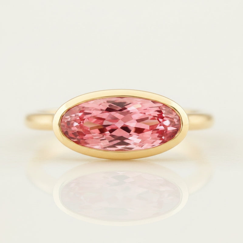An oval-cut pink gemstone ring set in a yellow gold bezel, displayed on a reflective surface with a soft reflection. The background is a soft beige gradient with a repeating watermark of 'MINIMAL ATELIER'.
