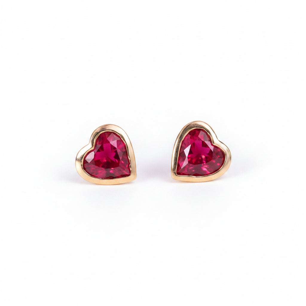 A pair of gold heart-shaped stud earrings with faceted red gemstones, displayed on a soft pink textured surface with a repeating 'MINIMAL ATELIER' watermark overlay.