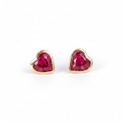 A pair of gold heart-shaped stud earrings with faceted red gemstones, displayed on a soft pink textured surface with a repeating 'MINIMAL ATELIER' watermark overlay.