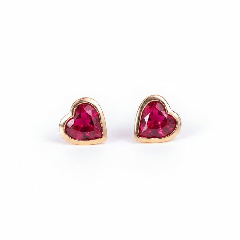 A pair of gold heart-shaped stud earrings with faceted red gemstones, displayed on a soft pink textured surface with a repeating 'MINIMAL ATELIER' watermark overlay.