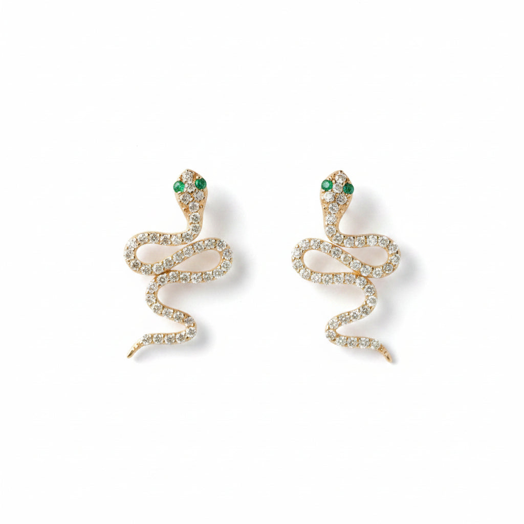 A pair of gold snake-shaped earrings adorned with clear rhinestones and small green gemstone eyes, displayed on a light textured background with a repeating 'MINIMAL ATELIER' watermark.