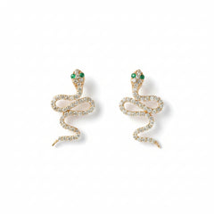 A pair of gold snake-shaped earrings adorned with clear rhinestones and small green gemstone eyes, displayed on a light textured background with a repeating 'MINIMAL ATELIER' watermark.