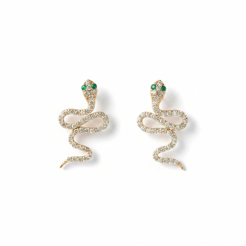 A pair of gold snake-shaped earrings adorned with clear rhinestones and small green gemstone eyes, displayed on a light textured background with a repeating 'MINIMAL ATELIER' watermark.