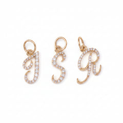 Three rose gold initial letter charms, 'J', 'S', and 'R', each paved with small clear stones, displayed on a dark textured background.