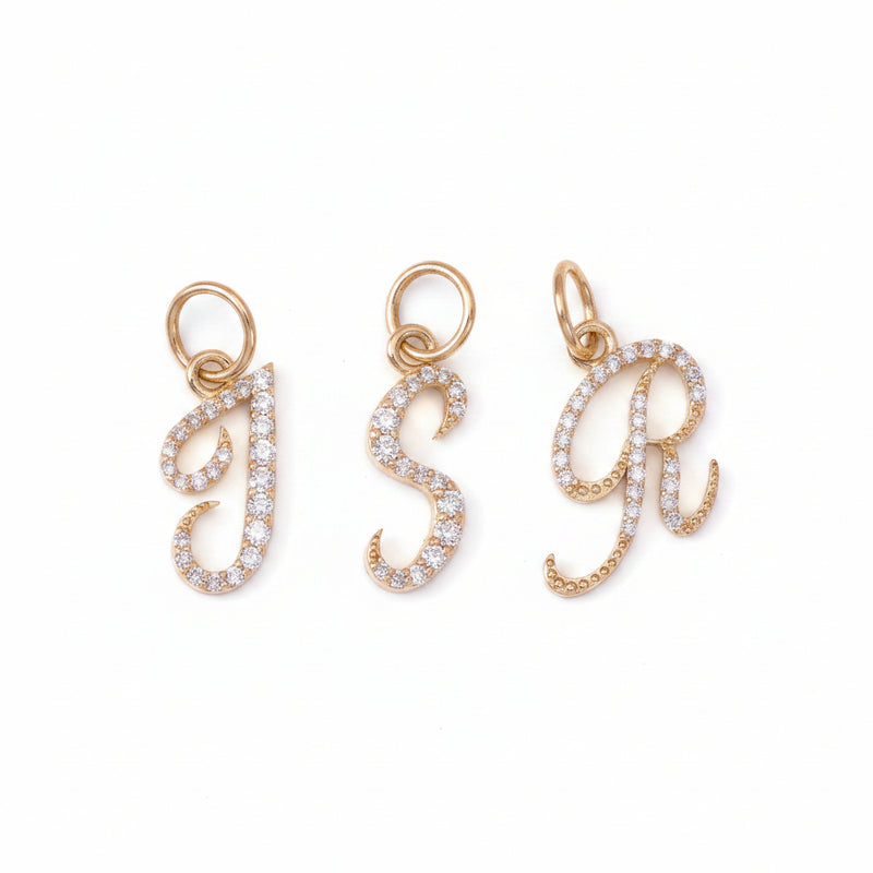 Three rose gold initial letter charms, 'J', 'S', and 'R', each paved with small clear stones, displayed on a dark textured background.