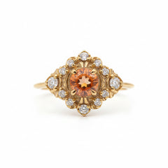A yellow gold ring featuring a prominent round orange gemstone in the center, surrounded by a halo of small clear stones, set against a light gray background with a repeating watermark.