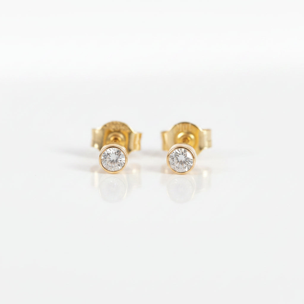 A pair of gold stud earrings with a single round clear gemstone in a bezel setting, displayed on a light pink background with a repeating 'MINIMAL ATELIER' watermark.
