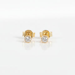 A pair of gold stud earrings with a single round clear gemstone in a bezel setting, displayed on a light pink background with a repeating 'MINIMAL ATELIER' watermark.