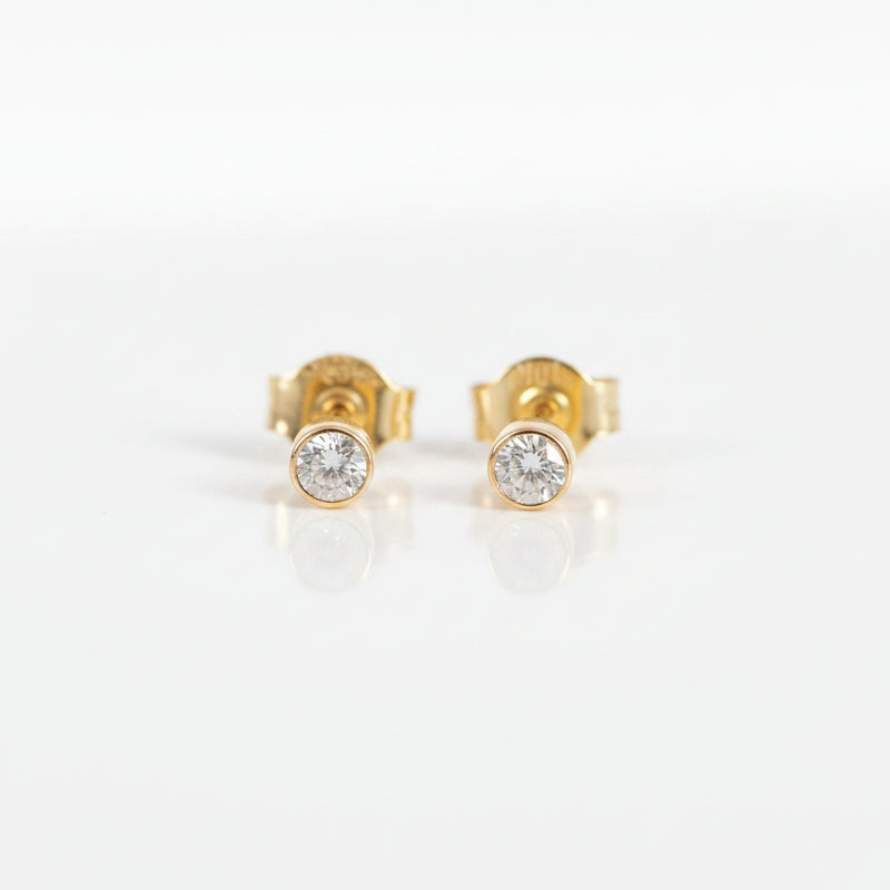 A pair of gold stud earrings with a single round clear gemstone in a bezel setting, displayed on a light pink background with a repeating 'MINIMAL ATELIER' watermark.