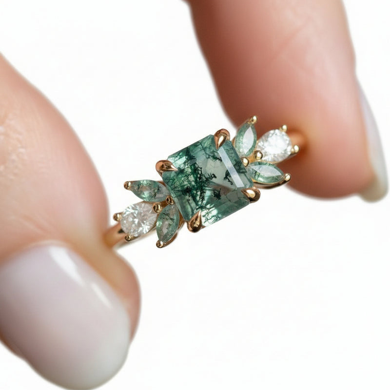 A close-up of a gold ring held between fingers, featuring a central green marbled stone with smaller clear stones on either side.