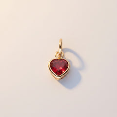 A gold heart-shaped charm with a red heart-shaped stone, shown on a light background with a repeating watermark pattern.