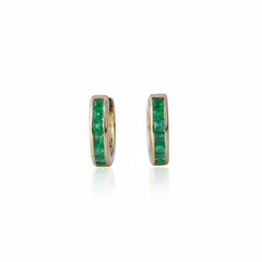 A pair of gold hoop earrings with a channel-set row of green emerald-cut stones, displayed on a light reflective surface.