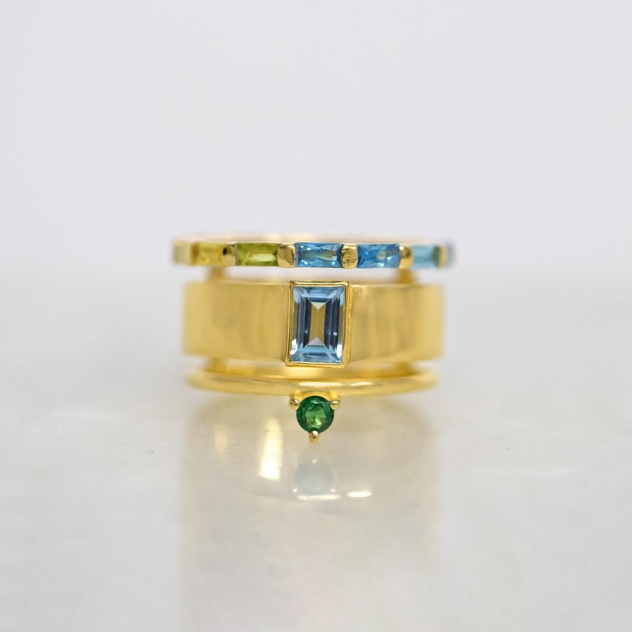 A gold ring with a central rectangular blue gemstone, flanked by a thin band with green and blue stones, and a smaller green gemstone dangling below, set against a light gray background.