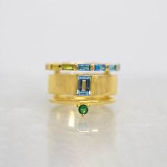 A gold ring with a central rectangular blue gemstone, flanked by a thin band with green and blue stones, and a smaller green gemstone dangling below, set against a light gray background.