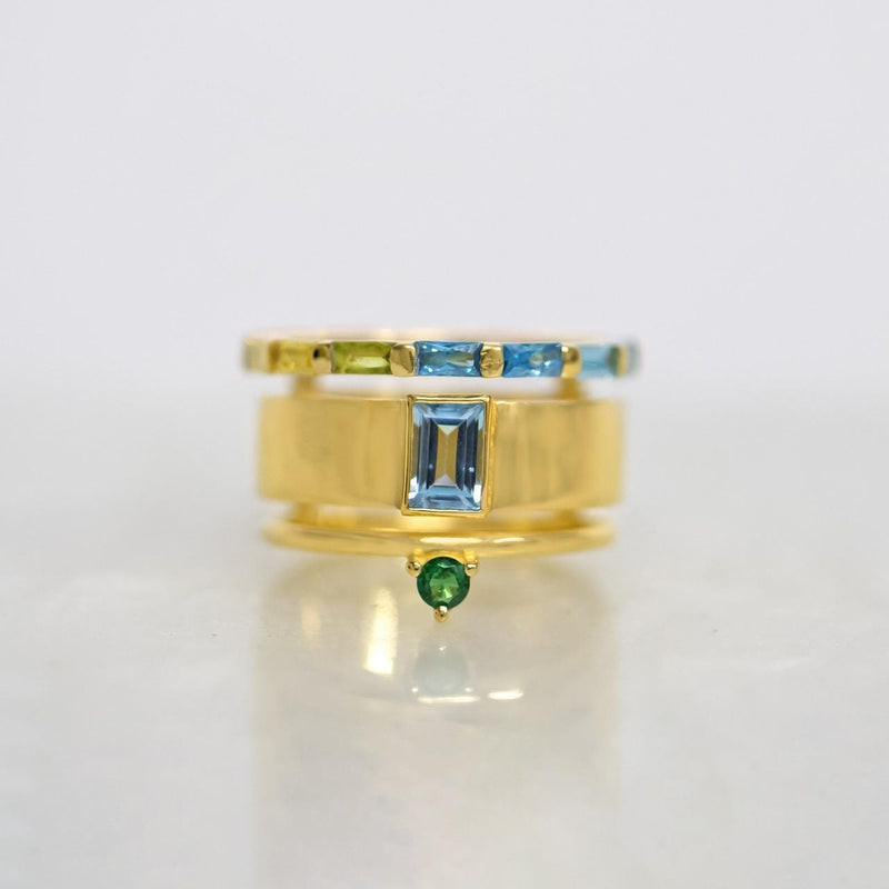 A gold ring with a central rectangular blue gemstone, flanked by a thin band with green and blue stones, and a smaller green gemstone dangling below, set against a light gray background.
