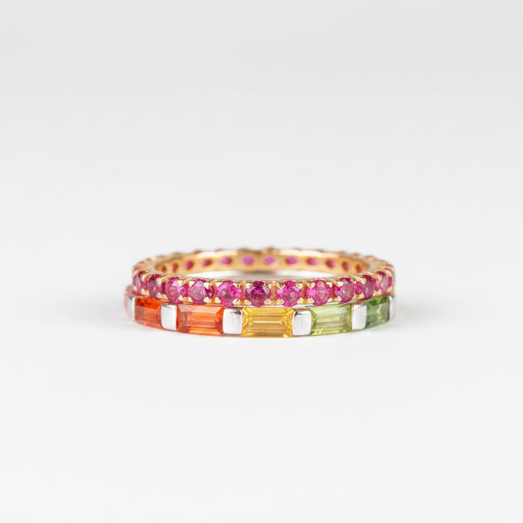 A close-up of a gold eternity ring featuring multiple rows of colorful gemstones, including pink, orange, yellow, and green, resting on a piece of natural wood against a neutral gray background.
