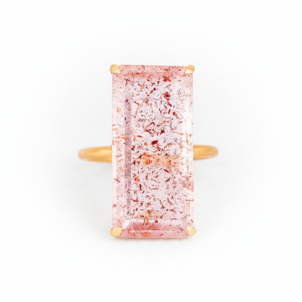 A rectangular pink gemstone ring with gold-toned metal prongs and a gold band, shown on a light gray textured background with a faint repeating watermark.