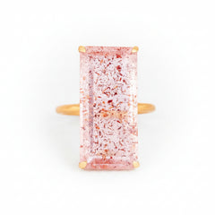 A rectangular pink gemstone ring with gold-toned metal prongs and a gold band, shown on a light gray textured background with a faint repeating watermark.