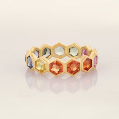 A gold honeycomb band ring featuring multiple faceted gemstones in various colors including orange, yellow, blue, and green, displayed on a light reflective surface with a faint reflection.