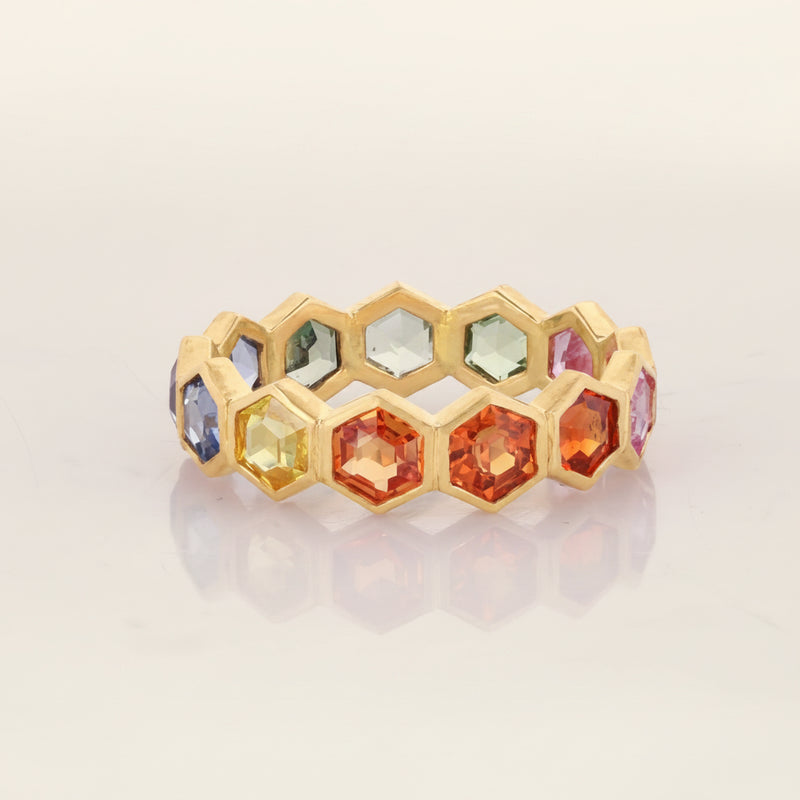 A gold honeycomb band ring featuring multiple faceted gemstones in various colors including orange, yellow, blue, and green, displayed on a light reflective surface with a faint reflection.