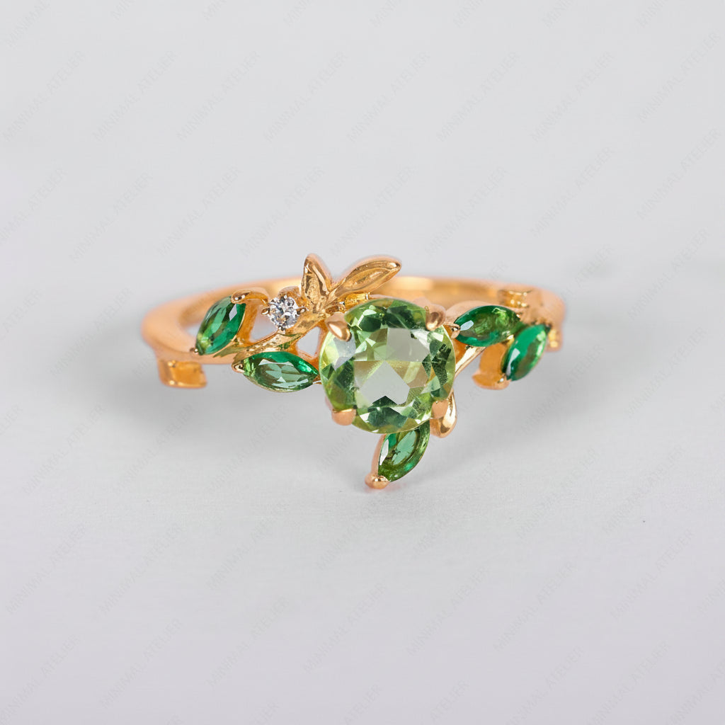 A gold ring designed as a leafy vine, featuring a large round green crystal centerpiece, smaller marquise-cut green crystals, and a tiny clear accent stone, shown on a light gray background.