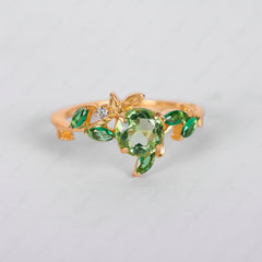 A gold ring designed as a leafy vine, featuring a large round green crystal centerpiece, smaller marquise-cut green crystals, and a tiny clear accent stone, shown on a light gray background.