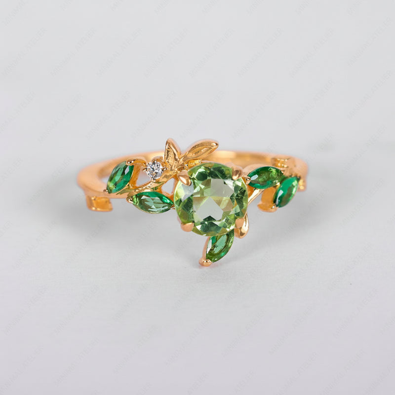 A gold ring designed as a leafy vine, featuring a large round green crystal centerpiece, smaller marquise-cut green crystals, and a tiny clear accent stone, shown on a light gray background.