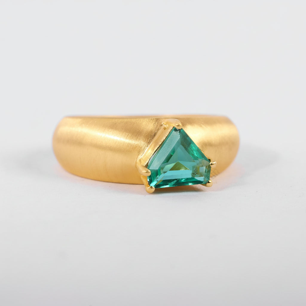 A gold ring with a triangular green gemstone in a prong setting, photographed on a light gray background with a repeating watermark.