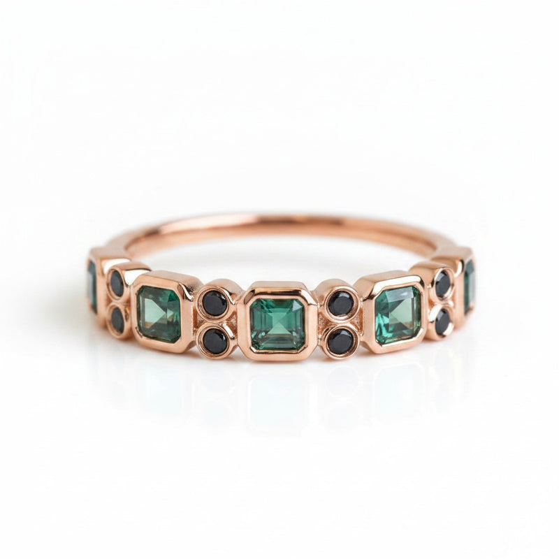 A rose gold eternity ring featuring square-cut green emerald-cut crystals and small round black gemstones, shown on a light gray reflective surface.