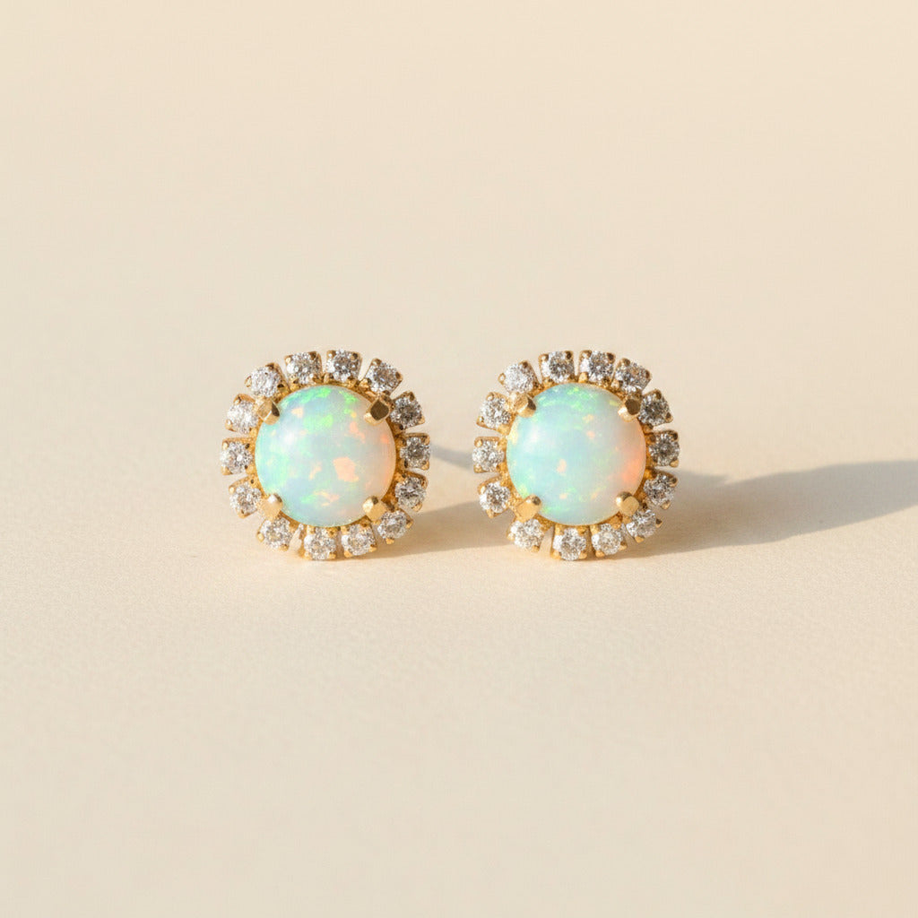 A pair of oval stud earrings with iridescent blue-green opal centers surrounded by small clear stones in a gold-tone setting, displayed on a textured gray surface.