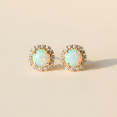 A pair of oval stud earrings with iridescent blue-green opal centers surrounded by small clear stones in a gold-tone setting, displayed on a textured gray surface.