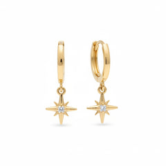 A pair of gold huggie hoop earrings with dangling starburst charms set with clear crystals, displayed on a textured beige surface.