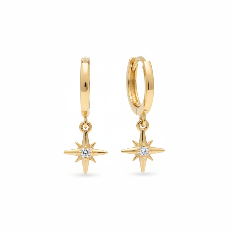 A pair of gold huggie hoop earrings with dangling starburst charms set with clear crystals, displayed on a textured beige surface.