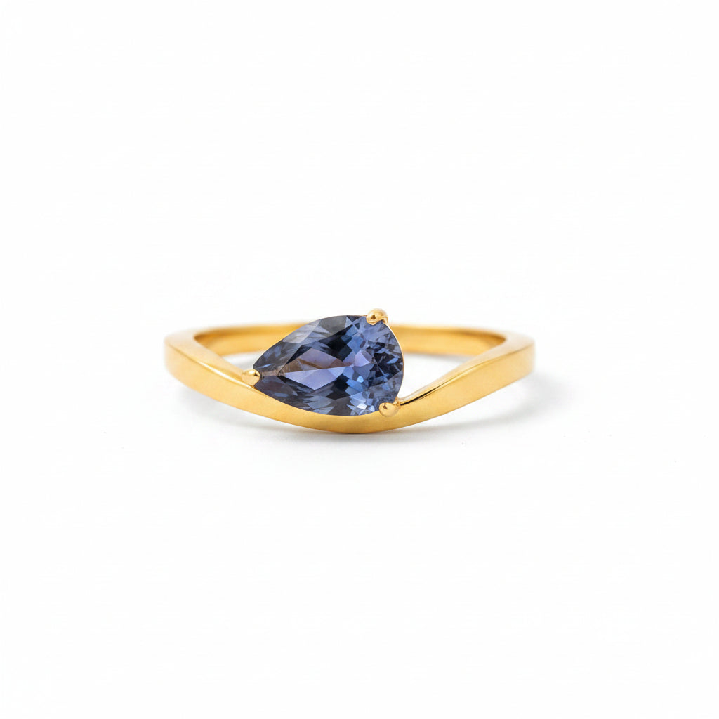 A gold ring with a teardrop-shaped blue gemstone resting on a white display block against a warm, blurred brown background.