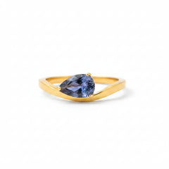 A gold ring with a teardrop-shaped blue gemstone resting on a white display block against a warm, blurred brown background.