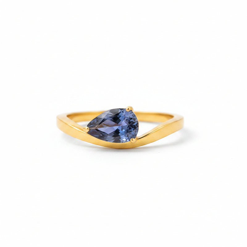 A gold ring with a teardrop-shaped blue gemstone resting on a white display block against a warm, blurred brown background.